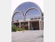 Facciata di un grande edificio moderno con ampie vetrate a griglia e copertura ad arco, caratterizzata da due grandi archi decorativi sovrapposti. Al piano terra si vedono un portico con pilastri, ingressi con cancelli e un piazzale asfaltato con aiuole.