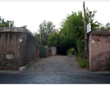 Ingresso a una strada stretta asfaltata, con pavimentazione in sampietrini in primo piano e due muri in cemento ai lati con numeri civici e segnaletica. Sullo sfondo la via prosegue tra vegetazione fitta e alberi, con cancelli e recinzioni lungo i lati.