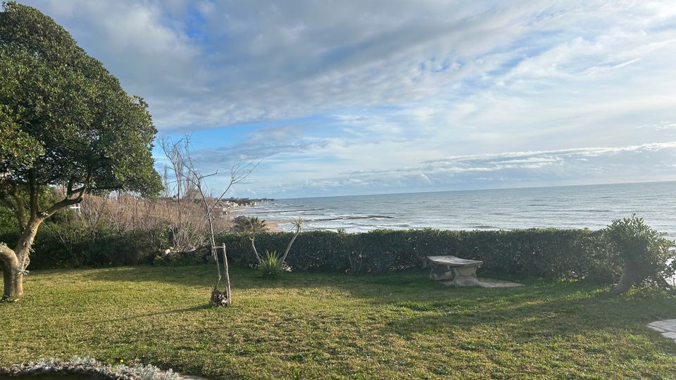 Giardino costiero con prato, siepi basse e alcuni alberi, affacciato su una spiaggia e sul mare con piccole onde. In primo piano si vede una panchina in pietra e un piccolo stagno delimitato da pietre, sotto un cielo parzialmente nuvoloso.