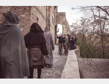 Stradina acciottolata lungo un edificio in pietra e mattoni con un arco e una lanterna, affiancata da un parapetto che si affaccia su alberi spogli in un paesaggio collinare. Sullo sfondo si vedono attrezzature cinematografiche posizionate sul percorso.