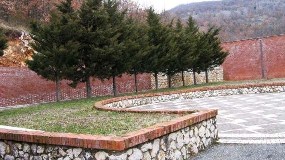 Cortile pavimentato con aiuole rialzate delimitate da muretti in pietra e bordo in mattoni, con una fila di alberi sempreverdi davanti a un alto muro in mattoni. Sullo sfondo si vedono colline boscose.