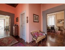 Interno di un appartamento con pareti rosa e pavimento in parquet, con una poltrona in vimini e quadri alle pareti. Due ampie aperture conducono a una zona pranzo con tavolo e sedie su tappeto e a un’altra stanza con tavolino e porta finestra.
