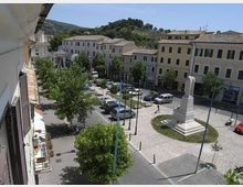 Veduta dall’alto di una piazza cittadina con alberi, parcheggi e una zona pedonale in ghiaia al centro. Nella piazza si trova un monumento in pietra su basamento, circondato da edifici storici a più piani con tetti in tegole e colline verdi sullo sfondo.