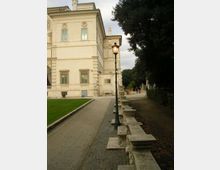 Viale pedonale accanto a un grande edificio storico in pietra chiara, con finestre alte e dettagli decorativi sulla facciata. Lungo il percorso ci sono un lampione in stile classico e una balaustra in pietra, con alberi fitti e area verde sul lato opposto.
