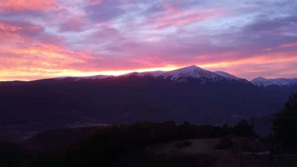 Panorama montano al tramonto con una lunga cresta in controluce e una cima parzialmente innevata sullo sfondo. Il cielo è attraversato da nuvole rosa e arancioni, con colline scure e vegetazione in primo piano.