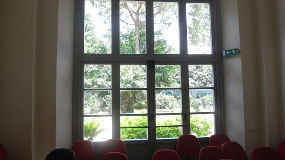 Sala interna con file di sedie imbottite rosse (e una nera) rivolte verso una grande porta-finestra a più riquadri. Attraverso i vetri si vede un’area verde con alberi e vegetazione, con segnaletica di uscita sulla parete a destra.