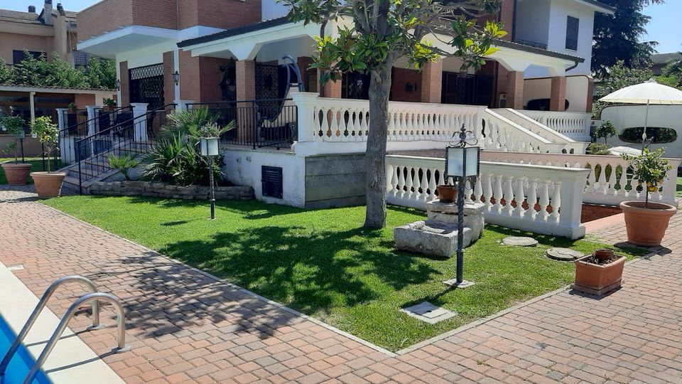 Villa a due piani in mattoni e intonaco bianco con portico e terrazza con balaustre, affacciata su un giardino con prato, albero centrale, lampioncini e vasi. In primo piano si vede un vialetto pavimentato e il bordo di una piscina.