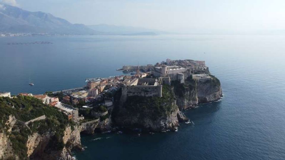 Veduta aerea di un promontorio roccioso sul mare con una città costiera compatta e una grande fortezza in pietra affacciata su acque calme. Sullo sfondo si intravedono montagne e una linea di costa lontana.