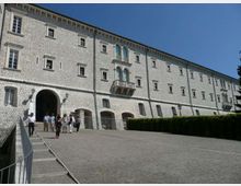 Lungo edificio storico in pietra chiara con facciata regolare di finestre e un grande arco d’ingresso, affacciato su un ampio viale in salita pavimentato. Al centro spicca una piccola loggia con balcone e finestre ad arco, con siepi lungo il bordo destro.