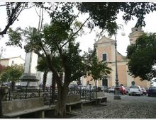 Piazza lastricata in pietra con alberi e panchine in primo piano, delimitata da una ringhiera in ferro e con un lampione. Sullo sfondo si vede una chiesa con facciata color pastello e campanile, affiancata da auto parcheggiate e da un monumento in pietra.