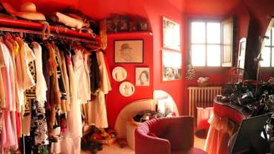 Interno di una stanza-guardaroba con pareti rosse, scaffalature e lunghi appendiabiti pieni di vestiti. Si vedono una finestra con termosifone sotto, una poltrona e diversi specchi, con borse e accessori appesi.