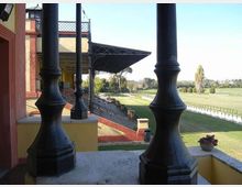 Vista da una tribuna coperta con colonne in ghisa e struttura metallica, affacciata su una pista erbosa recintata e ampi campi verdi. A sinistra si vede l’edificio della tribuna con pareti color ocra e rosso e gradinate sottostanti.