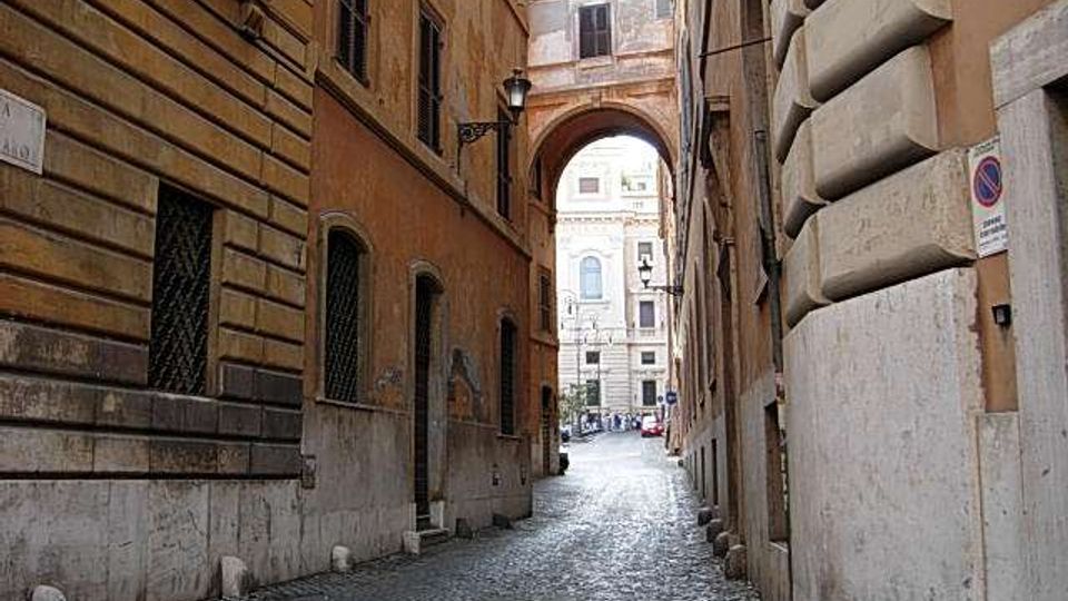 Stretto vicolo urbano in pietra con pavimentazione a sampietrini, fiancheggiato da alti edifici storici in muratura. In fondo si apre un arco che conduce verso una strada più ampia con facciate monumentali visibili oltre il passaggio.