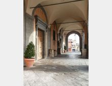 Portico in pietra con pavimentazione in lastre e serie di arcate a volta; lungo il lato sinistro si vedono porte incorniciate e vasi con piante. In fondo, un grande arco apre sulla strada esterna con edifici visibili oltre il passaggio.