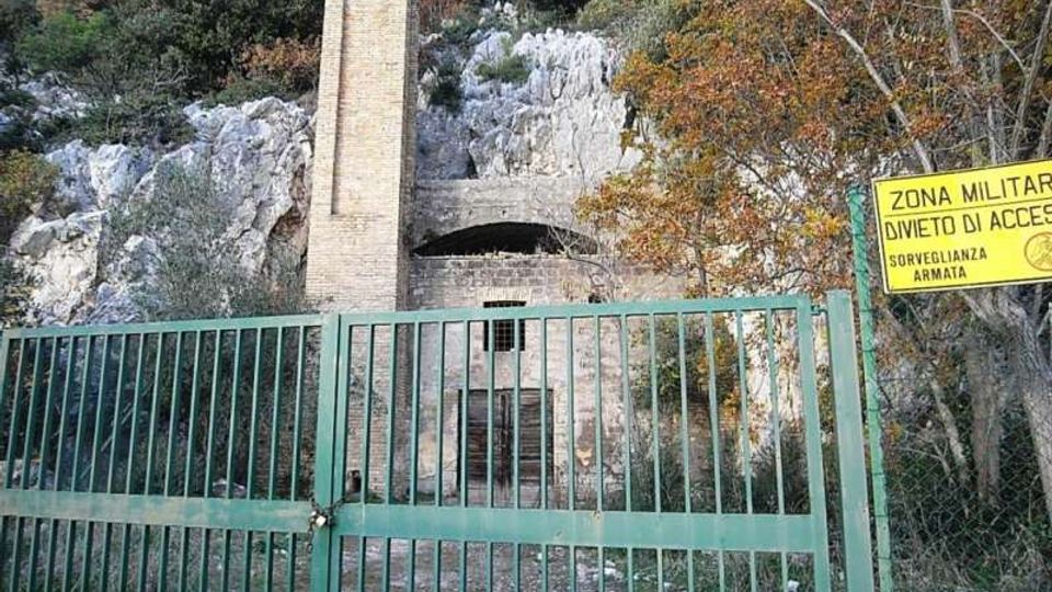Cancello metallico verde che chiude l’accesso a una struttura in pietra incassata nella parete rocciosa, con alto camino in mattoni e portone murato. A destra è visibile un cartello giallo “Zona militare, divieto di accesso”, circondato da vegetazione e alberi.