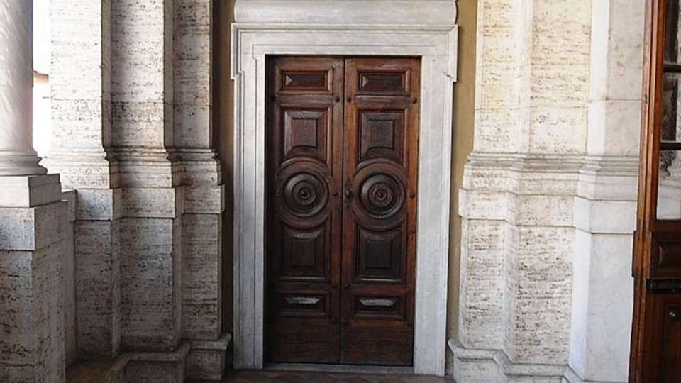 Ingresso interno di un edificio storico con grandi colonne in pietra e una porta doppia in legno scuro incorniciata da una cornice chiara. In primo piano si vede un pavimento a mosaico geometrico con motivi a rombi e bordure decorative.
