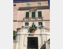 Facciata di un edificio storico a più piani con intonaco chiaro, finestre con persiane verdi e cornici decorative. Al centro si vede un balcone semicircolare con balaustra, vaso e ombrellone chiuso sopra un grande portone con inferriata in ferro battuto.