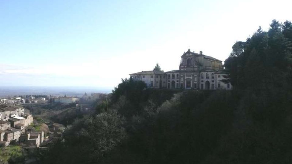 Veduta panoramica di un complesso religioso con chiesa e edifici adiacenti su un colle, affacciato su una valle. In primo piano si vedono pendii alberati, mentre a sinistra si estende un piccolo centro abitato con case in pietra.