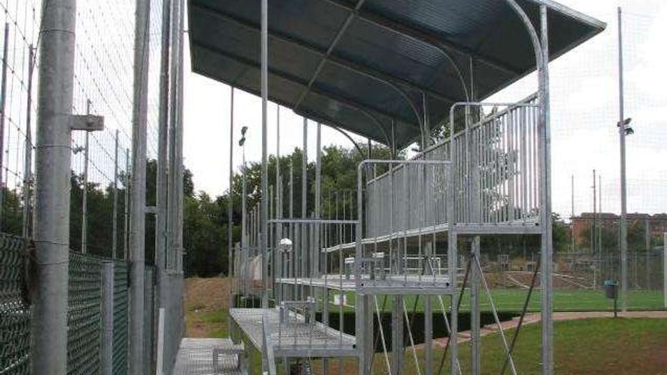 Piccola tribuna metallica con gradinate e corrimano, coperta da una tettoia inclinata, all’interno di un impianto sportivo all’aperto. La struttura è affiancata da alte recinzioni a rete e circondata da prato e terreno battuto, con alberi sullo sfondo.