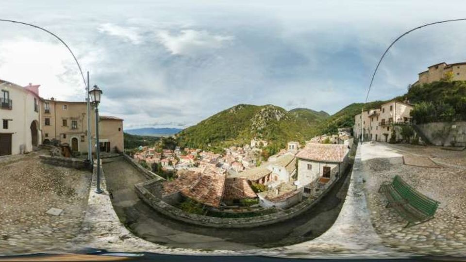 Veduta panoramica da una terrazza belvedere in pietra con pavimentazione a ciottoli, lampione e panchina, affacciata su un borgo di case in pietra con tetti in coppi. Sullo sfondo si vedono colline verdi e un castello o fortificazione sulla sommità, sotto un cielo nuvoloso.