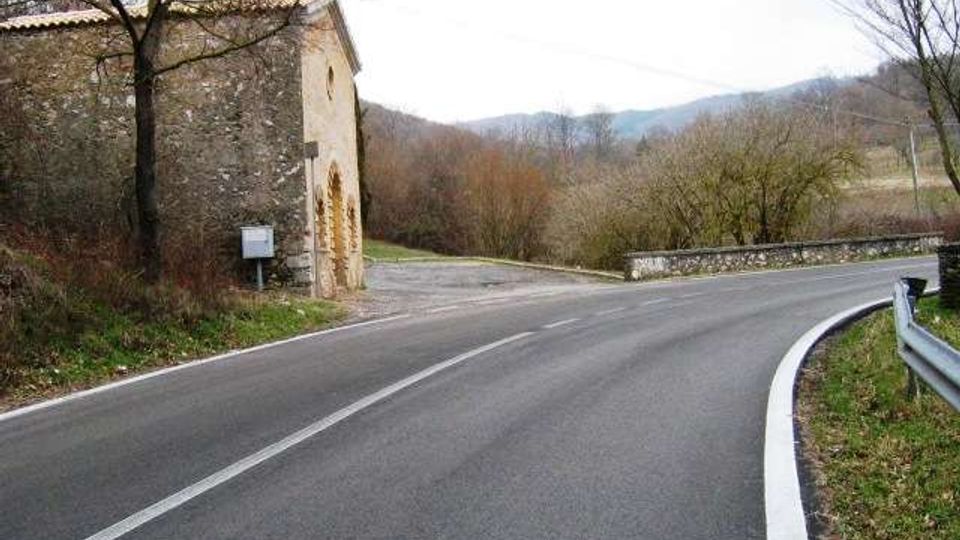 Strada asfaltata di campagna che curva a destra, con guardrail metallico e segnaletica a bordo carreggiata. A sinistra si vede un edificio in pietra vicino al margine della strada, con alberi spogli e colline sullo sfondo.