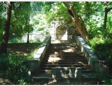 Scalinata in pietra in un parco alberato, con parapetti laterali e vegetazione fitta ai lati. In cima ai gradini si vede una recinzione metallica e, sullo sfondo, un arco in muratura parzialmente illuminato tra gli alberi.