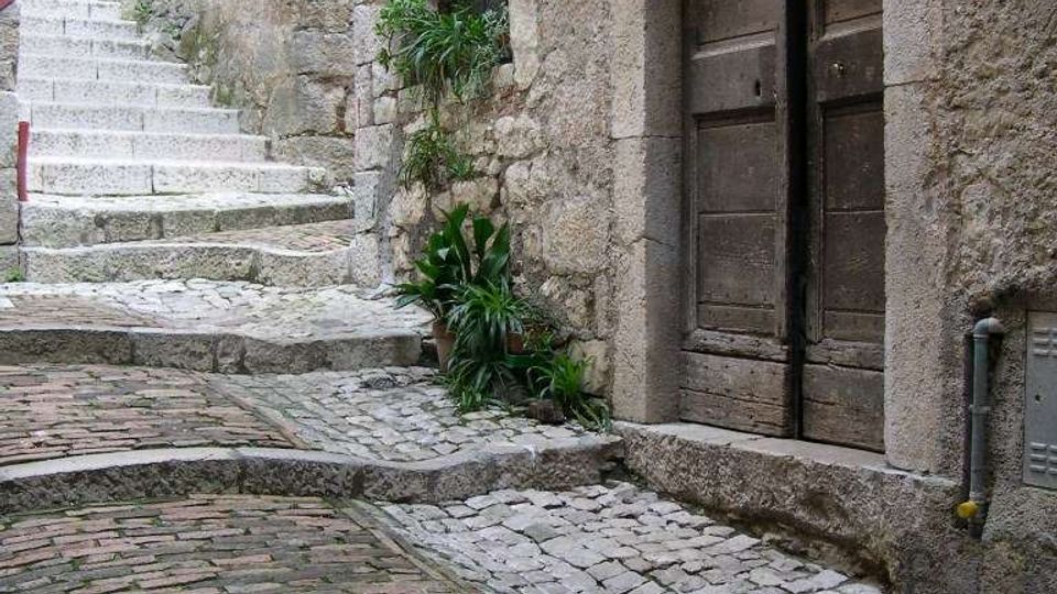 Vicolo in pietra acciottolata in un borgo storico, con pavimentazione a gradoni che sale lungo muri in muratura grezza. A destra si vede un grande portale ad arco con doppia porta in legno, affiancato da piccole piante e una finestra incassata.