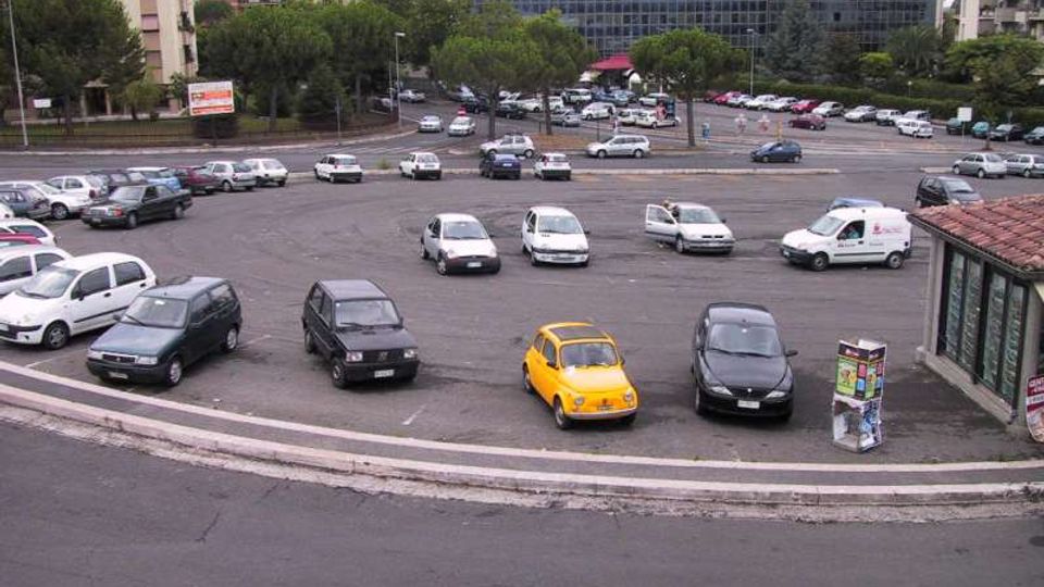 Ampia rotatoria adibita a parcheggio, con numerose auto disposte lungo la curva e carreggiata asfaltata in primo piano. Sullo sfondo si vedono alberi, edifici residenziali e un grande edificio moderno con facciata in vetro, oltre a un’area con auto parcheggiate lungo la strada.