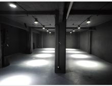 Ampio spazio interno tipo magazzino o sala industriale vuota, con pareti scure, pavimento in cemento e colonne centrali che sostengono il soffitto basso. Lampade a sospensione con cavi a vista illuminano a chiazze il pavimento lungo il corridoio centrale.