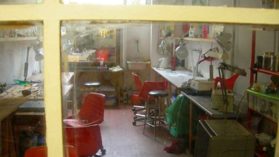 Vista attraverso una finestra a riquadri di un piccolo laboratorio artigianale con tavoli da lavoro, lampade da banco e utensili. Nella stanza si vedono diverse sedie rosse e scaffali con materiali e contenitori lungo le pareti.