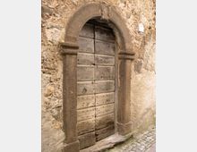 Porta antica in legno a due ante, con chiodature e battente in ferro, inserita in un arco in pietra scolpita su un muro di muratura grezza. Davanti alla soglia si vede un tratto di pavimentazione in ciottoli lungo una stretta strada.