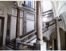 Atrio interno di un edificio storico con grandi colonne in pietra e archi a volta decorati. Una scalinata in marmo con balaustre conduce ai piani superiori illuminati da ampie finestre.