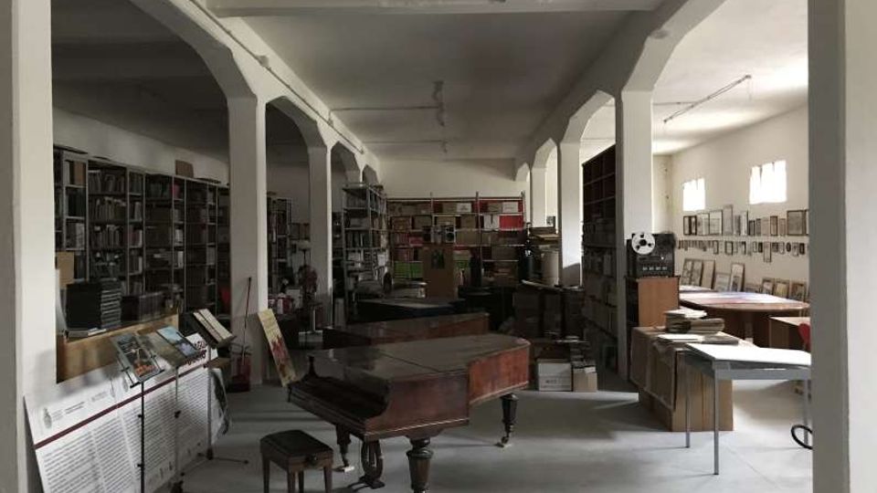 Ampia sala interna tipo archivio o laboratorio, con colonne bianche che sostengono il soffitto e scaffalature piene di libri e materiali lungo le pareti. Al centro ci sono un pianoforte a coda e tavoli da lavoro, con finestre sulla destra che illuminano l’ambiente.