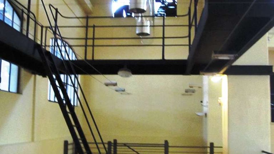 Interno di un edificio industriale riconvertito con soffitto alto, soppalco in metallo con parapetti e scala inclinata. Grandi finestre, pavimento in legno e condotte di ventilazione a vista sul soffitto.