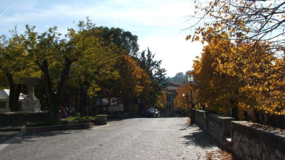 Strada in pavé leggermente in discesa, fiancheggiata da alberi con foglie autunnali e da un muretto in pietra sul lato destro. Sullo sfondo si vedono lampioni e edifici tra la vegetazione sotto un cielo limpido.