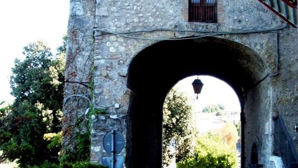 Passaggio ad arco sotto un edificio in pietra con facciata irregolare, finestra con balconcino e una lanterna sospesa al centro della volta. La strada acciottolata attraversa l’arco e conduce verso un’area con alberi e auto parcheggiate, con muretto laterale e cavi a vista sulla parete.