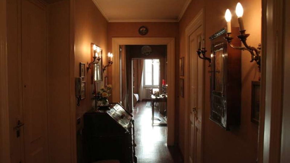 Corridoio interno di un appartamento d’epoca con pareti color ocra, pavimento in legno e applique a parete in stile classico. Ai lati si vedono porte e piccoli arredi con specchi e quadri, mentre in fondo una finestra illumina una stanza adiacente.