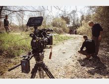Sentiero sterrato e ghiaioso in un’area boschiva con vegetazione rada e alberi spogli, attraversato da un piccolo corso d’acqua poco profondo. In primo piano è presente una videocamera professionale su treppiede, con attrezzatura di ripresa lungo il bordo del sentiero.