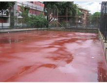 Campo sportivo all’aperto con pavimentazione rossa bagnata dalla pioggia e pozzanghere, circondato da alte recinzioni metalliche. Sullo sfondo si vedono alberi e edifici residenziali e industriali ai margini dell’area.