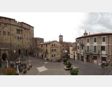 Piazza lastricata in un centro storico, circondata da edifici in pietra con persiane e portici, e dominata a sinistra da un grande edificio fortificato medievale con scalinata. Sullo sfondo si vede una torre con orologio e al centro della piazza sono presenti fioriere e arredi urbani.