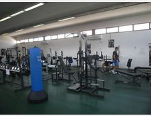 Interno di una palestra con soffitto alto e finestre a nastro, pavimento verde e pareti bianche. Sono visibili panche, rastrelliere per pesi, macchine da allenamento e un sacco da boxe blu al centro della sala.