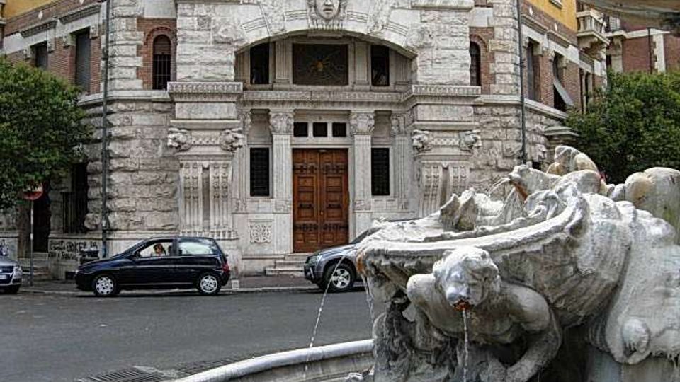 Fontana in pietra con una figura scolpita che versa acqua in una vasca semicircolare, in primo piano lungo una strada urbana. Sullo sfondo si vede la facciata di un edificio storico in muratura e mattoni, con ingresso ad arco e portone in legno.