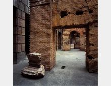 Interno di rovine in mattoni con grandi archi e passaggi, con pareti antiche segnate da fori e aperture. Pavimento in cemento e un blocco di pietra spezzato in primo piano, affiancati da una parete moderna rivestita in lastre scure.