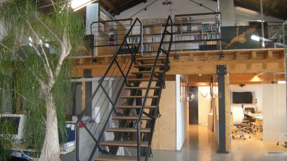 Interno di un loft adibito a ufficio con soppalco in legno e scala centrale in metallo, soffitto alto con travi a vista e lucernari. Sul soppalco ci sono scaffali con libri e una ringhiera metallica, mentre al piano terra si vedono area riunioni e una grande pianta in vaso.