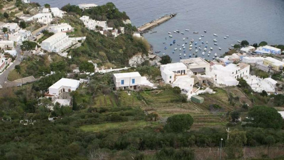Veduta dall’alto di un villaggio costiero con case bianche su un pendio verde terrazzato, affacciato su una piccola baia. In mare si vede un molo e numerose barche ancorate vicino alla riva.