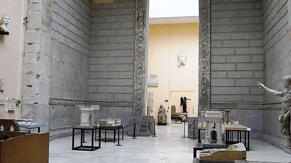 Interno di un museo con ampie pareti in pietra e pavimento chiaro, organizzato come galleria con modelli architettonici su tavoli e vetrine. Al centro si vede un grande portale monumentale in pietra decorata che conduce a una sala successiva.