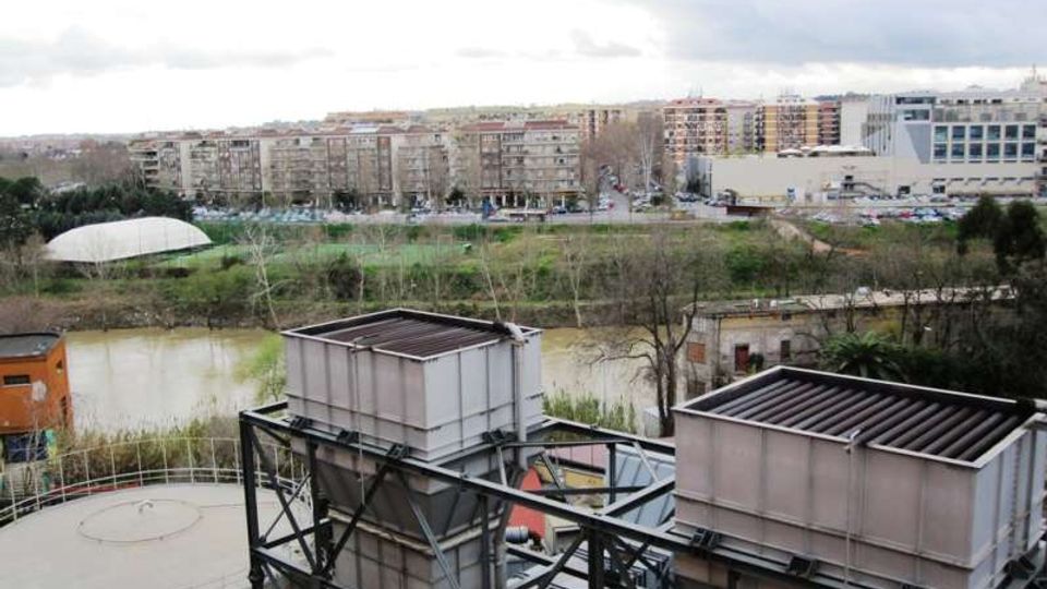 Vista urbana con un fiume in primo piano e, davanti, grandi strutture industriali metalliche e un serbatoio circolare. Sullo sfondo si vedono aree verdi, una strada e palazzi residenziali e commerciali sotto un cielo nuvoloso.