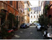 Vicolo urbano stretto con pavimentazione in sampietrini, fiancheggiato da edifici alti con facciate color ocra e persiane. Auto e scooter parcheggiati lungo i lati, piante in vaso e panni stesi su fili tesi tra i palazzi in fondo alla strada.