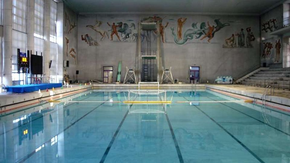 Interno di una piscina coperta con vasca rettangolare e corsie segnate sul fondo, circondata da bordo in piastrelle e attrezzature da nuoto. Sulle pareti alte sono presenti grandi murales con figure atletiche, gradinate laterali e ampie finestre che illuminano l’ambiente.