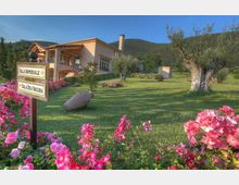 Giardino curato con prato verde e aiuole di fiori rosa, ulivi e un grande anfora decorativa, con un edificio moderno a due piani sullo sfondo e colline boscose dietro. In primo piano un cartello direzionale indica “Sala Imperiale” e “Sala dei Vacuna”.